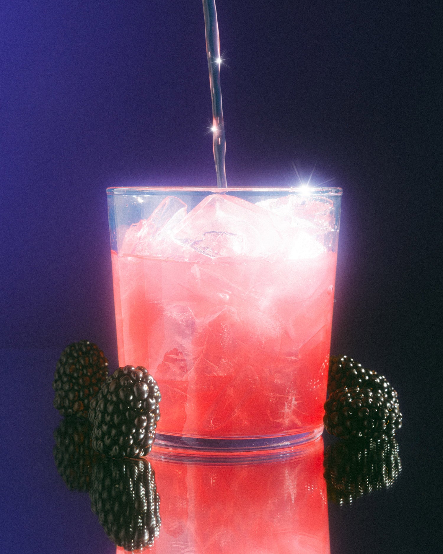 Red cocktail with ice being poured into a glass against a dark background