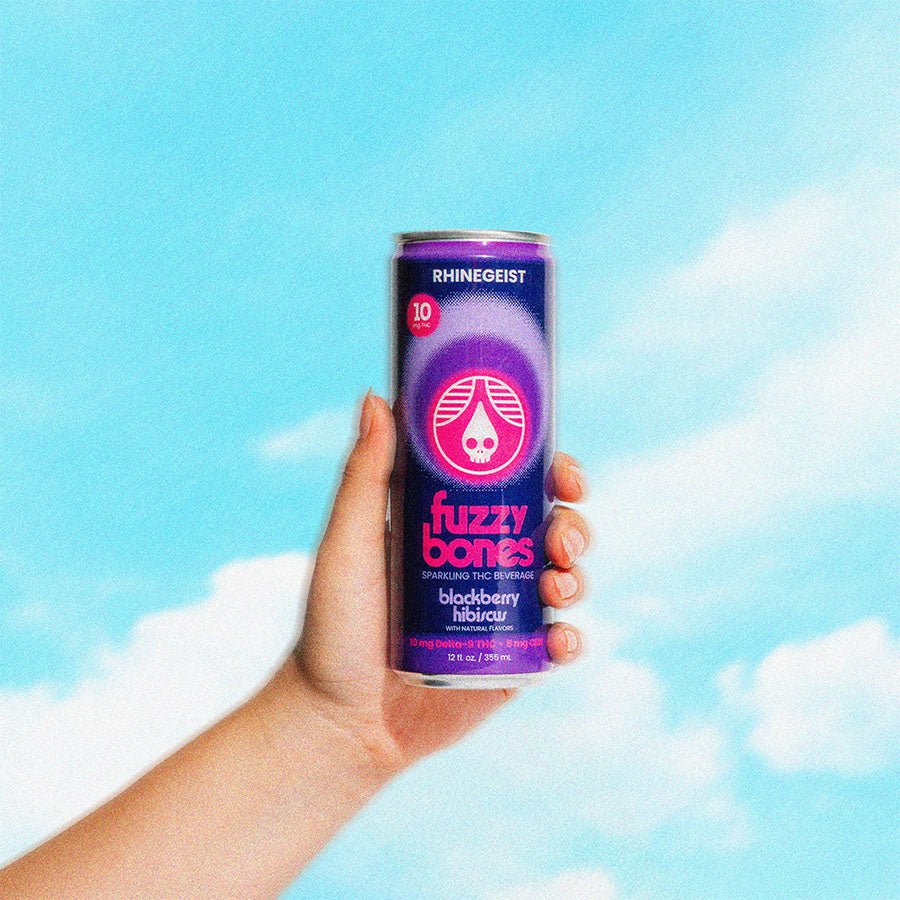 Hand holding a can of Fuzzy Bones blackberry hibiscus against a blue sky with clouds